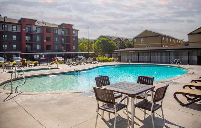 A pool with chairs and a table in front of apartment buildings.