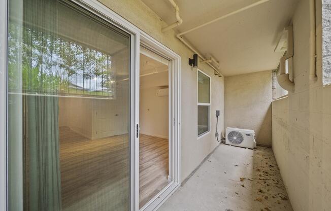 View of a small balcony or patio area with sliding glass doors leading to an interior room. The space features a light-colored wall, an air conditioning unit, and a floor with scattered leaves. Natural light filters through the doors, reflecting on the interior wooden flooring.