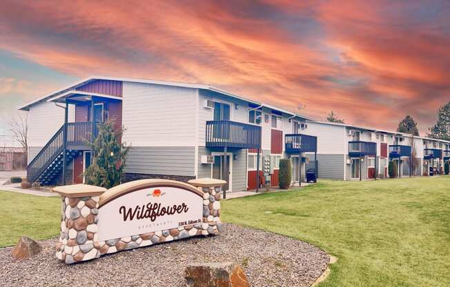Outside the community with a Wildflower Apartments sign, apartments in the background surrounded by green grass, landscaping, and a colorful sunset.at Wildflower, Kennewick