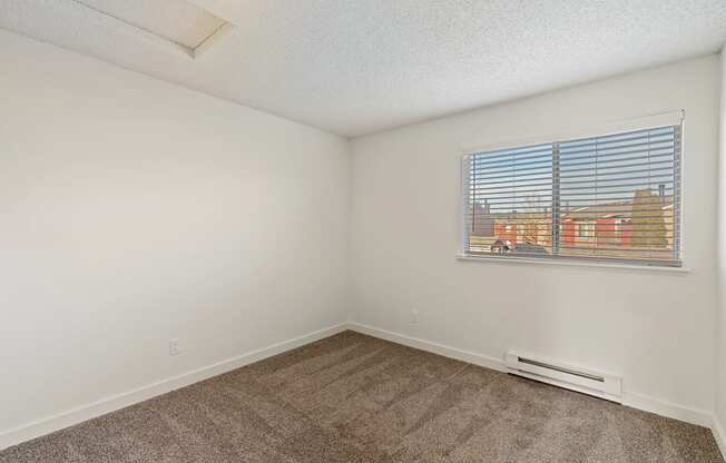 A bedroom with white walls, taupe carpets, a double window with white blinds, and a view on a sunny day.