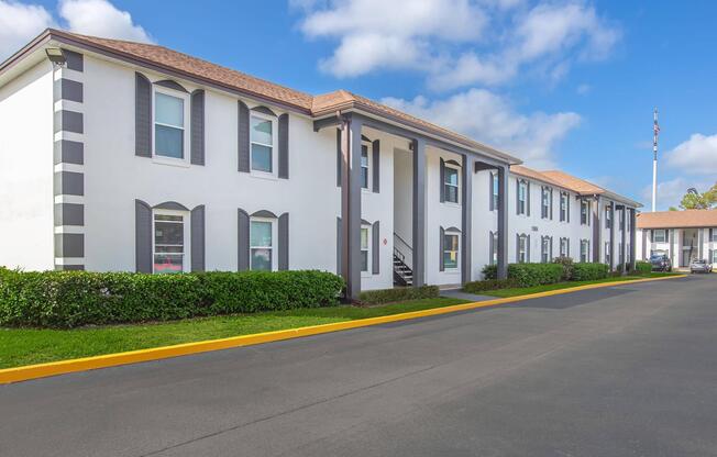 A two-story building with a white exterior and dark gray accents, featuring large windows and a staircase. The foreground includes a neatly landscaped area with shrubs, and the road is paved. Blue skies with scattered clouds are visible above the structure.
