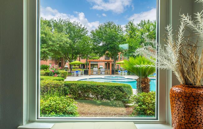 View from a window overlooking a pool area surrounded by lush greenery and umbrellas. The scene features palm trees, flower beds, and a cozy outdoor seating area, creating a relaxing atmosphere under a partly cloudy sky.