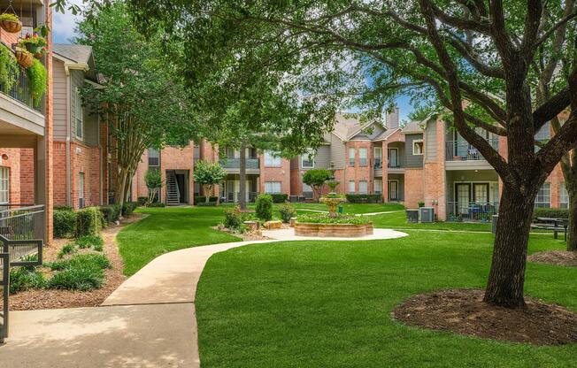 A landscaped courtyard of an apartment complex featuring manicured lawns, a stone fountain, and pathways. Surrounding the courtyard are brick buildings with balconies and greenery, creating a serene outdoor space for residents. Bright, sunny skies add to the ambiance.