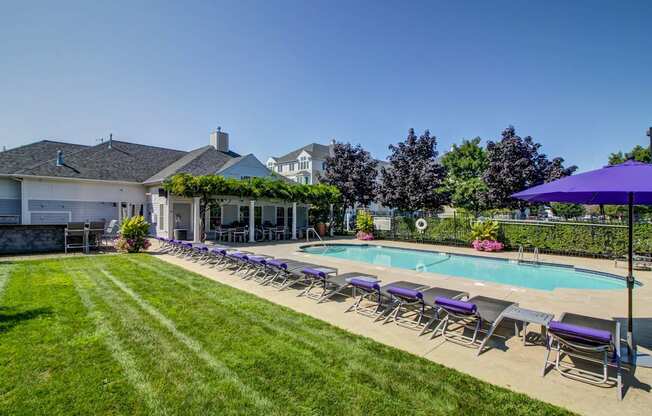 View of pool with purple lounge chairs and lush green grass surrounding. Clubhouse with greenery covered deck can be seen in background.