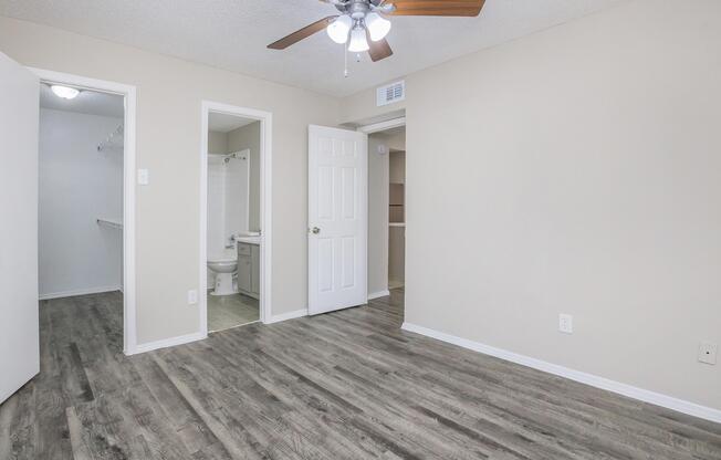 A spacious, well-lit room with wood-look flooring, a ceiling fan, and a light-colored wall. Doors leading to a closet and a bathroom are visible. The overall ambiance is clean and inviting, suitable for a bedroom or living space.