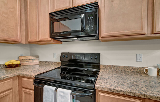 A modern kitchen here at Swan Creek with warm wood cabinets, black electric range and microwave, and spacious granite countertops, creating a clean, functional, and welcoming cooking space.