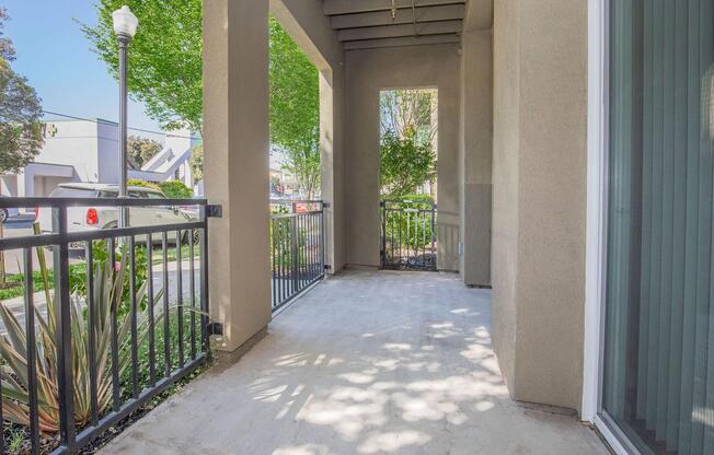 A concrete porch with a railing, bordered by green plants. Sunlight casts shadows on the ground, and leafy trees can be seen beyond the railing. The area is well-lit and offers a view of the surroundings, creating a welcoming outdoor space.