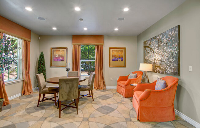 A comfortable resident lounge here at Asbury Park featuring a round dining table with upholstered chairs, two plush accent armchairs with a side table and lamp, patterned carpet flooring, framed wall art, soft neutral walls, ceiling lighting, and large windows with drapery that bring in natural light and create a relaxed gathering space.