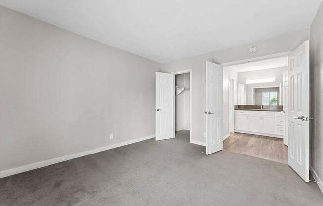 A room with white walls and carpeted floor with a kitchen visible through an open door.