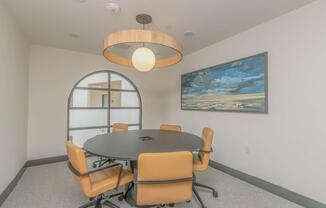 A modern conference room featuring a round black table surrounded by five orange chairs. An artwork depicting a seascape hangs on the wall, and a large circular light fixture is suspended from the ceiling. An arched window adds a contemporary touch to the space.