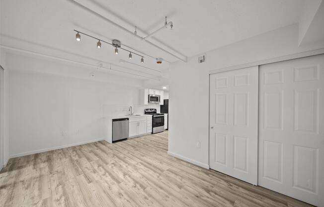 A modern, open-concept living space featuring light wood flooring, white walls, and recessed lighting. The kitchen is equipped with black and stainless steel appliances, including a microwave and stove. There are dual sliding closet doors on the right, enhancing the room's functionality and minimalistic design.