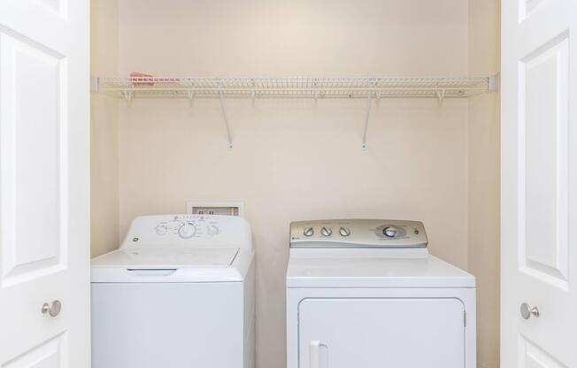 A laundry room featuring a white washing machine and a white dryer side by side, with a wire shelf above them. The walls are painted beige, and the entrance is framed by two closed white doors. The space is clean and organized, perfect for laundry tasks.