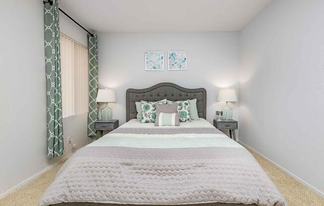 A neatly made bed with a gray upholstered headboard in a softly lit bedroom. The bedding features a light gray and white pattern. Two bedside tables with lamps are on either side of the bed. The walls are painted light gray, and there are decorative curtains and two framed artworks above the bed.