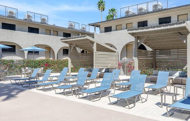 A sunlit outdoor pool area featuring multiple blue lounge chairs arranged neatly. There are shaded cabanas and umbrellas, with greenery in the background and modern buildings in view. The setting suggests a relaxing and inviting atmosphere near a pool.