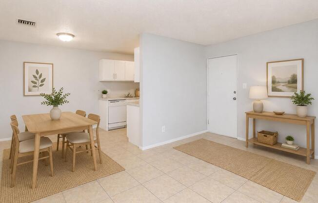 A bright and airy kitchen and dining area featuring a wooden dining table with four chairs, light-colored walls, and tiled flooring. The kitchen is partially visible with white cabinetry and appliances. Two framed botanical prints decorate the walls, and a console table with decorative items is positioned near the entrance.