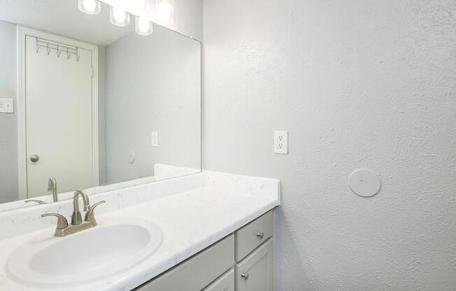 A clean and simple bathroom featuring a white marble countertop with a sink, modern faucet, and vertical mirror. The walls are painted a light gray, and there is a door leading to another room. The space is well-lit with a light fixture above the sink.