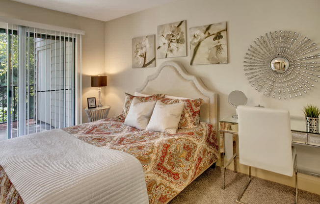 A comfortable bedroom here at Heritage Hills featuring a plush upholstered bed dressed in patterned bedding and layered pillows, soft neutral walls with coordinated floral artwork, and carpeted flooring. The space includes a mirrored desk with a modern chair, a decorative sunburst mirror, bedside lighting, and sliding glass doors that open to a private balcony with leafy views.