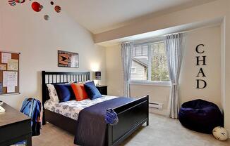 Three-Bedroom Apartments in Kenmore, WA - Northshore Townhomes - Bedroom with Carpeted Flooring, Bed, Nightstand with Lamp, Desk Chair, Bean Bag, Vaulted Ceiling, and Large Window.