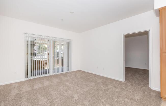 Spacious interior of a room with beige carpet, featuring large windows with blinds and a doorway leading to another room. The walls are painted white, creating a bright and airy atmosphere. No furniture is present, emphasizing the open space.