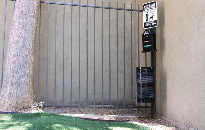 Pasadena Brookmore apartments dog run with artificial turf, a tall tree, and a pet waste disposal station with trash bags and trash can.