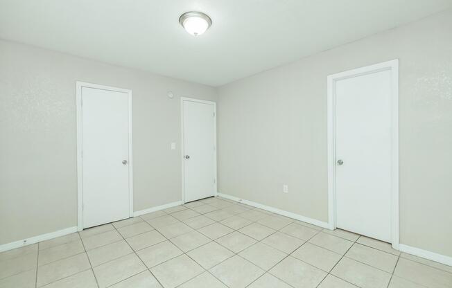 A plain room with light gray walls and tiled flooring. There are three white doors on the walls, two of which are on the left and one on the right. The ceiling features a simple light fixture, and the overall space appears empty and unadorned, creating a minimalist atmosphere.