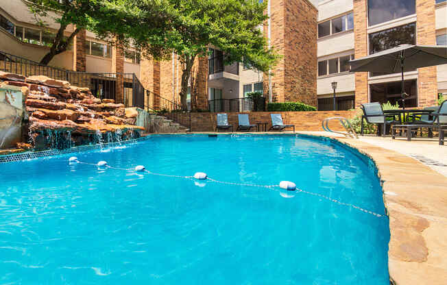 A swimming pool with a waterfall surrounded by lounge chairs and a couple of tables, chairs and umbrellas at Cambridge Court Apartments Entryway in Lake Highlands, Dallas, TX.