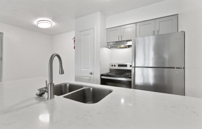A modern kitchen with a white marble countertop featuring a double sink. The background includes sleek gray cabinets, a stainless steel refrigerator, and an electric stove with an oven. Bright overhead lighting illuminates the space, emphasizing the clean and contemporary design.