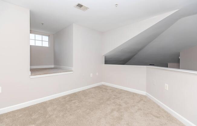 Open space in a light-colored room with beige carpet. One wall features a sloped ceiling, and there's a small window allowing natural light. A short staircase leads to a landing area. The overall environment appears clean and modern, suitable for various uses, such as a living area or office.