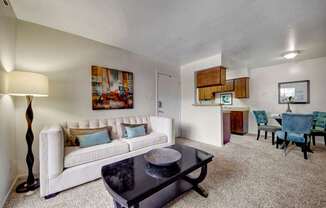 A bright living room here at Northridge Court showcasing a comfortable sofa with accent pillows, a sleek coffee table on plush carpeting, and soft neutral walls, opening into a dining area with upholstered chairs and a kitchen pass-through that creates an open, inviting layout filled with natural light.