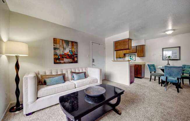 A bright living room here at Northridge Court showcasing a comfortable sofa with accent pillows, a sleek coffee table on plush carpeting, and soft neutral walls, opening into a dining area with upholstered chairs and a kitchen pass-through that creates an open, inviting layout filled with natural light.