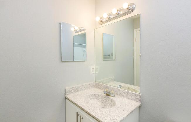 A clean and modern bathroom with a light-colored wall, featuring a granite countertop sink, a rectangular mirror above the sink, and a four-bulb light fixture. The space has a minimalistic design, with a white cabinet base and a door partially visible in the background.