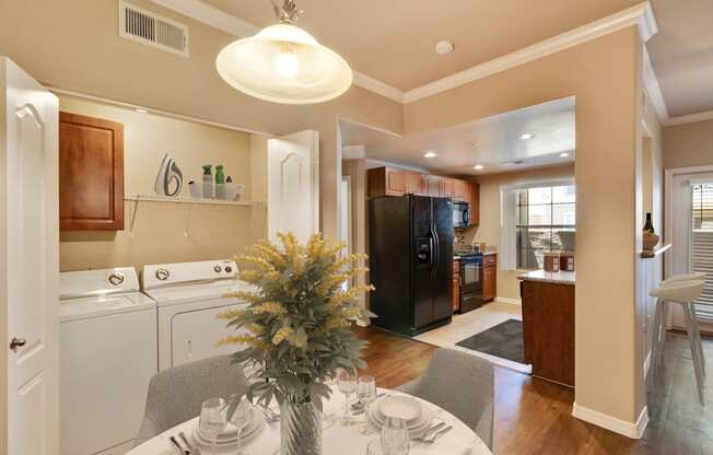 Photo of the kitchen from the dining area. The round dining table with 2 chairs can be seen. A bouquet of yellow flowers in a vase  sits on the dining table. A washer and dryer can be seen in a closet to the left with a folding door open showing the appliances can be hidden from view. A gloss black fridge and black stove can be seen in the kitchen.