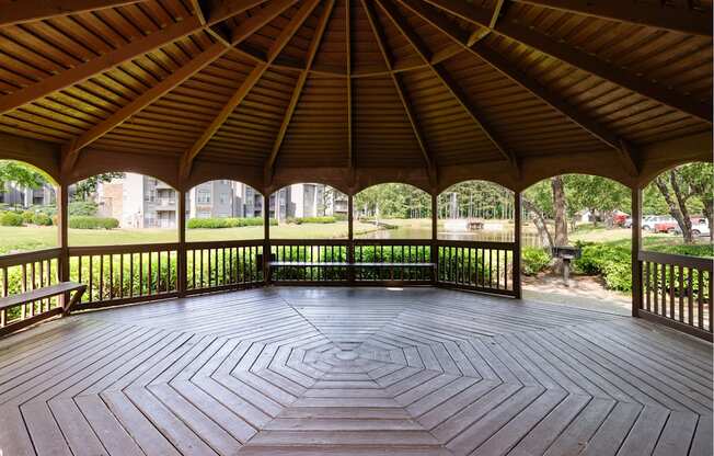 A wooden pavilion with a circular floor and a roof next to a pond and grill