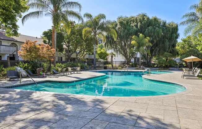 A large outdoor swimming pool surrounded by trees and a patio area with chairs.