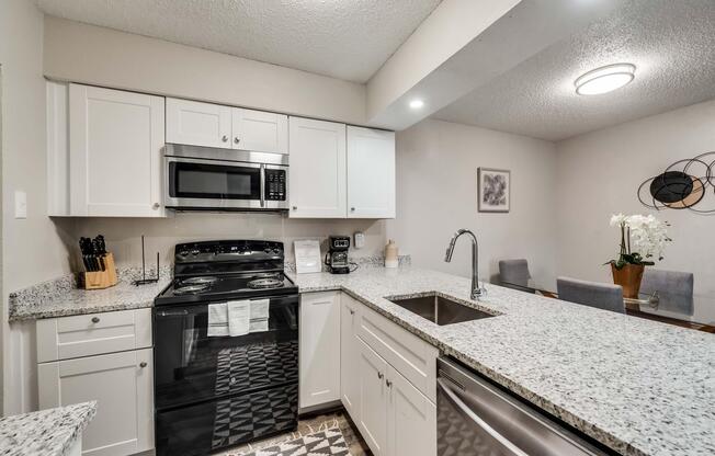 Modern kitchen with white cabinets, a black stove, and microwave. Countertops are covered in light gray speckled granite. Stainless steel sink with a sleek faucet, and a dining area in the background featuring a table with gray chairs and a decorative plant. Bright lighting enhances the space.