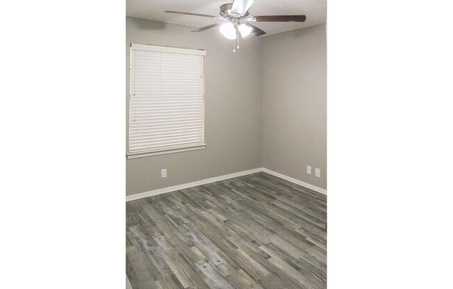 Empty room featuring light gray walls and a wooden floor. A ceiling fan with at least three blades is installed, and there's a window with white blinds. The space is well-lit and appears ready for furnishings, highlighting a clean and simple aesthetic.