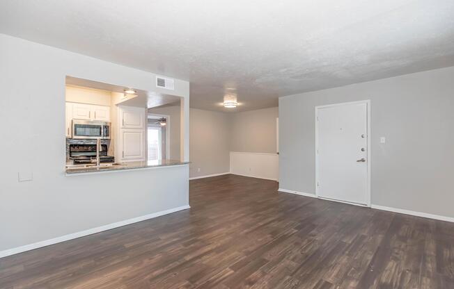 A spacious, modern living area with light gray walls and new wood laminate flooring. The open layout features a kitchen with a breakfast bar visible through an archway. A front door leads to the entrance, and there's a window letting in natural light. The space is clean and bright, ready for interior design.