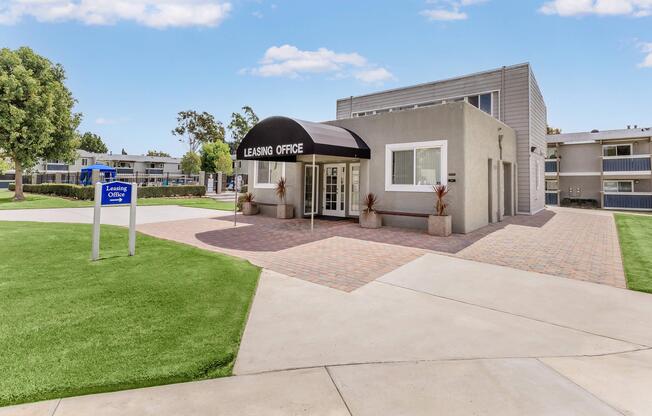 A modern leasing office with a black awning labeled "Leasing Office." The building features a gray exterior and is surrounded by well-maintained grassy areas and pathways. A sign stands nearby indicating the leasing office location.