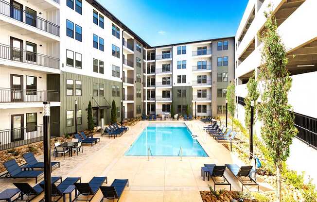A swimming pool surrounded by lounge chairs and trees in front of apartment buildings.