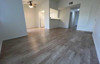 Interior view of a spacious, well-lit living area featuring a light-colored laminate floor, a ceiling fan, and an open layout leading to a kitchen with white cabinetry. The walls are painted in a neutral tone, enhancing the airy feel of the space.