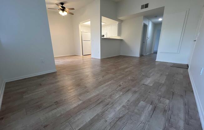 Interior view of a spacious, well-lit living area featuring a light-colored laminate floor, a ceiling fan, and an open layout leading to a kitchen with white cabinetry. The walls are painted in a neutral tone, enhancing the airy feel of the space.