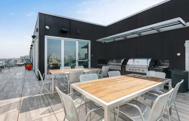 A modern rooftop grilling area here at Blue and Lime featuring stainless steel grills set beneath a sleek canopy and surrounded by wood-top dining tables with metal chairs. Wide plank decking extends across the terrace, with glass doors leading into an indoor lounge. Open sky views and ample seating create a functional space for outdoor cooking and gatherings.