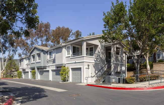 A multi-unit residential building with a gray exterior and a staircase leading to the entrance. The property features several garages, surrounded by light landscaping and trees, under a clear blue sky. The driveway is paved with a red curb marking the edge.