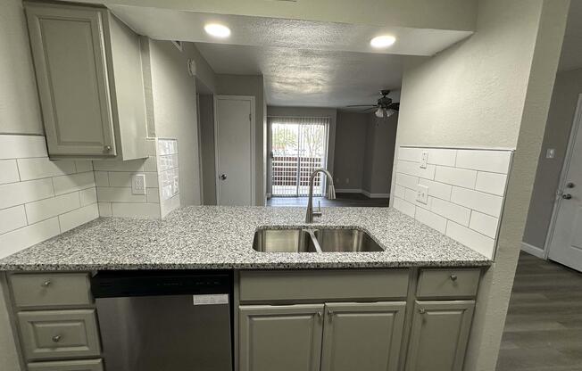 A modern kitchen featuring grey cabinetry, a granite countertop with a double sink, and stainless steel appliances. The background shows a living area with sliding doors leading to a balcony, illuminated by natural light. The walls are painted in neutral tones, creating a fresh and inviting atmosphere.
