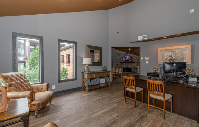 A modern lobby area featuring a brown leather armchair, a wooden side table with a lamp, and a reception desk with a computer. The walls are painted gray, and there are large windows allowing natural light in. Decor includes wall art and a cozy ambiance, with a view of an outdoor space through the windows.