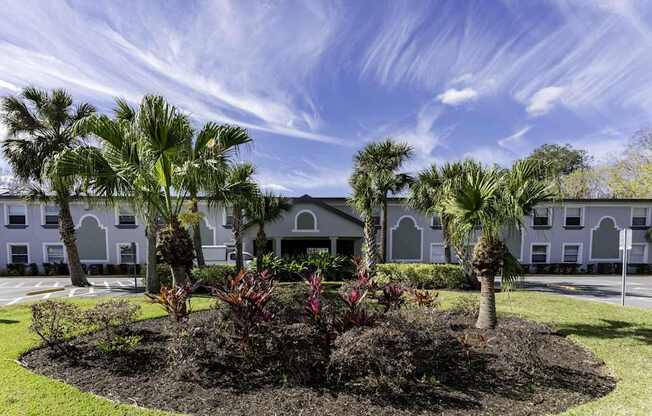 A white building with arched windows is surrounded by a well-manicured garden with palm trees.