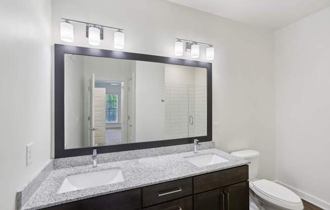 A bathroom with granite countertops, two sink, mirror, stand up glass panel shower, and toilet.