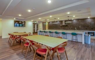 A modern communal kitchen and dining area featuring a large wooden table with chairs, green bar stools at a kitchen counter, and sleek appliances. The space has warm lighting, a TV mounted on the wall, and a minimalist design with wood flooring.