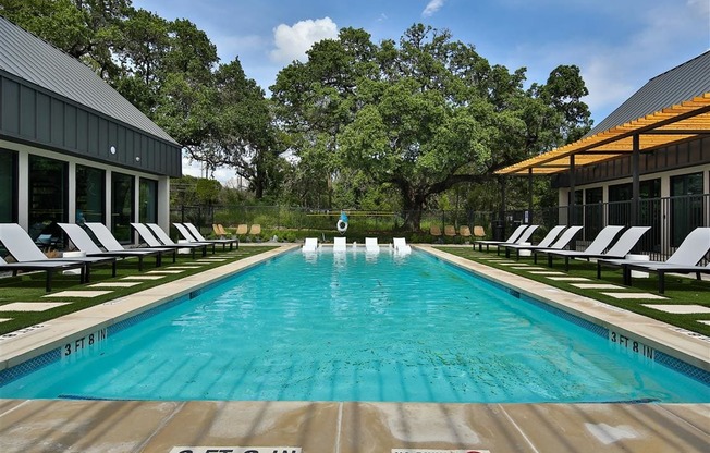 A gated swimming pool lined with lounge chairs near a clubhouse and large trees.