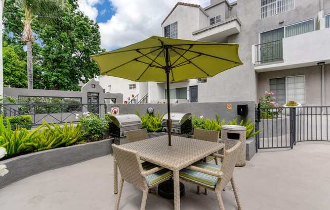 Outdoor table with chairs and lime green shade umbrella. Green shrubs in planter around the seating area and gas BBQ's.